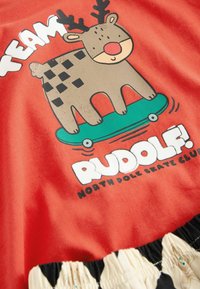 Rød bomulds t-shirt med et grafik af et rensdyr på en skateboard, med sorte pletmønstre og teksten "TEAM RUDOLPH! NORTH POLE SKATE CLUB."