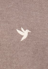 Geborduurde witte kolibrie op lichtbruin gebreid stof, met gedetailleerde steken en textuur.