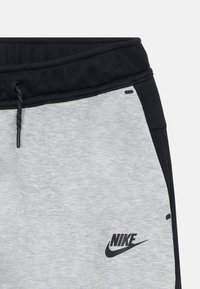 Pelēki un melni Nike bikses ar melnu šnouri, sānu kabatām un melnu logotipu apakšējā labajā stūrī. Mīksta, teksturēta auduma.