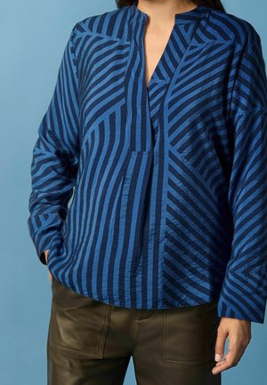 Personne portant une chemise à manches longues rayée bleue avec un col mao et un pantalon en cuir brun foncé, devant un fond bleu uni.