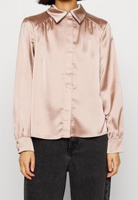 Chemise en satin rose clair à manches longues avec col pointu, fermeture à boutons et boutons aux poignets, présentant une texture lisse et un design structuré.