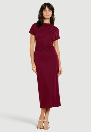 Femme debout portant une robe midi bordeaux ajustée à manches courtes avec un détail froncé, associée à des sandales dorées à bout ouvert sur un fond uni clair.