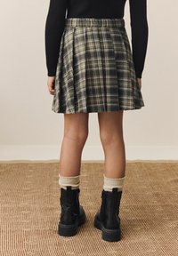Geruite rok in donker groen, zwart en wit; geplooid ontwerp. Draag met zwarte enkellaarsjes en crèmekleurige sokken. Effen achtergrond.