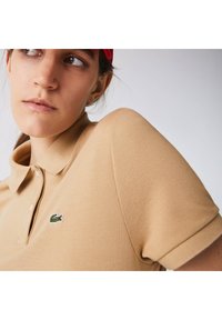 Beige polo-paita, jossa on teksturoitu kangas, lyhyet hihat ja pieni vihreä krokotiililogo rintaan. Klassinen kaulus.