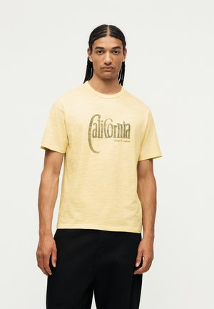 Jeune homme aux longues tresses portant un t-shirt jaune avec le texte « California Keep it rockin' », associé à un pantalon noir, en position neutre.