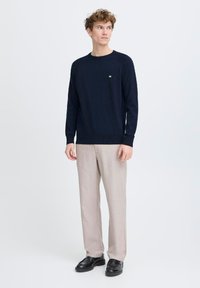 Jersey de cuello redondo en color navy, hecho de un suave tejido de punto, combinado con pantalones beige claros y zapatos negros. Presenta un pequeño logo en el pecho.
