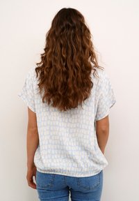 Chemisier bleu clair à manches courtes avec un motif texturé de formes beige clair. Coupe décontractée avec un ourlet froncé. Associé à un jean ajusté.