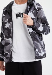 Leichte Nylonjacke in schwarzem und grauem abstraktem Muster. Mit Reißverschluss vorne, verstellbarer Kapuze und elastischen Bündchen. Wird über einem weißen T-Shirt getragen.