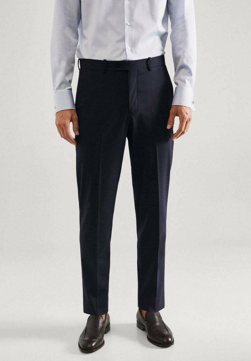 Pantalons de dress navy en tissu lisse, avec une coupe ajustée, des passants de ceinture et une légère tapered au niveau des chevilles ; associés à des mocassins noirs.
