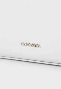 Geantă albă cu o suprafață netedă, logo-ul argintiu "Calvin Klein" și linii curate fără modele sau embellishments suplimentare.