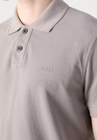 Polo gris clair avec deux boutons foncés et le logo "BOSS" sur la poitrine gauche, porté par une personne à la peau claire.