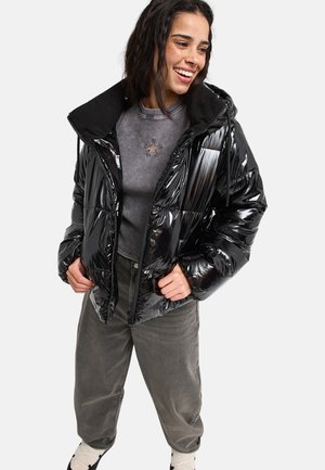 Frau lächelnd, trägt eine glänzende schwarze Steppjacke, grauen Rippsweater mit Stern-Detail, lockere graue Hose und karierten Socken mit Schuhen.