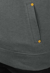 Sudadera gris con dobladillo acanalado, que cuenta con un bolsillo lateral y un botón de acento amarillo. La textura parece suave y informal.