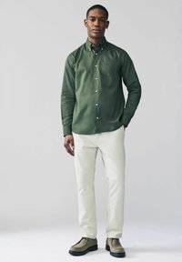 Camisa de botones verde oscuro con un bolsillo en el pecho, combinada con pantalones de un color claro y zapatos marrones, presentando una textura de tela suave.