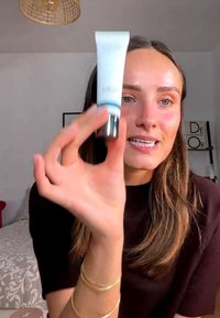 Frau mit langen braunen Haaren, die drinnen eine kleine weiße und silberne Kosmetiktube hält und präsentiert.