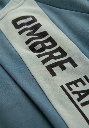 Lichtblauwe stof met een gladde textuur toont een opvallende zwarte grafiek met de tekst "OMBRE EAT." Het ontwerp bevat paneeldetails in contrasterende kleuren.