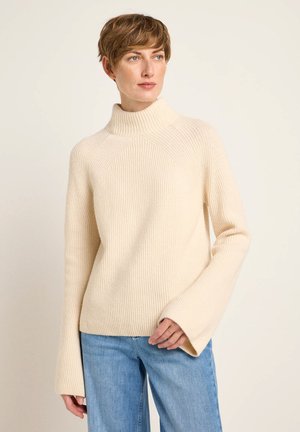 Cremfarbener gerippter Rollkragenpullover mit lockerer Passform und weit geschnittenen Ärmeln, der ein strukturiertes Strickdesign und eine einheitliche Farbe aufweist. Kombiniert mit blauen Jeans.