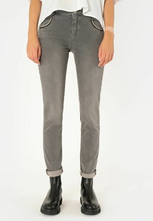 Osoba v šedých úzkých džínách s ohrnutými lemy a černých kotníkových botách, stojící před bílým pozadím.