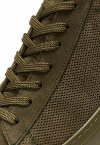 Olivgrüne Wildledersneaker mit gewebten Schnüren, strukturierten Perforationen an der Seite, kontrastierenden Nähten und einer Gummisohle.