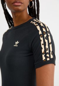 Kvinna som bär en svart Adidas-tröja med leopardmönstrade tre ränder på axlarna och en liten logotyp på bröstet, flätat hår och ringörhängen.