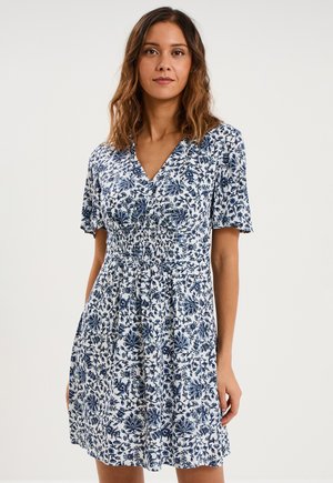 Femme aux cheveux bruns mi-longs portant une robe blanche à motifs floraux bleus et une taille froncée, debout devant un fond uni.