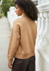 Bruine, oversized sweatshirt met een hoge hals en geribde manchetten, gecombineerd met glanzende donkerbruine broeken, tegen een witte reling achtergrond.