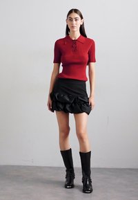 Haut rouge à côtes à manches courtes avec un col et trois boutons, associé à une mini-jupe noire en satin à volants. Des bottes noires montantes complètent le look.