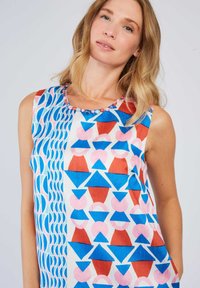 Frau mit blonden Haaren, die ein ärmelloses Kleid mit blauen, roten und pinken geometrischen Formen sowie einem bestickten Ausschnitt trägt, vor einem einfarbigen Hintergrund.
