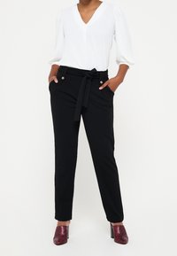 Černé kalhoty na míru s opaskem na zavazování, předními kapsami a jemnými zlatými knoflíky, doplněné bílou blůzkou s výstřihem do V a strukturovanými červenými lodičkami.