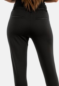Pantalones negros ajustados de corte slim con un tejido texturizado que presenta pequeños lunares dorados. Los bolsillos traseros están sutilmente integrados en el diseño.