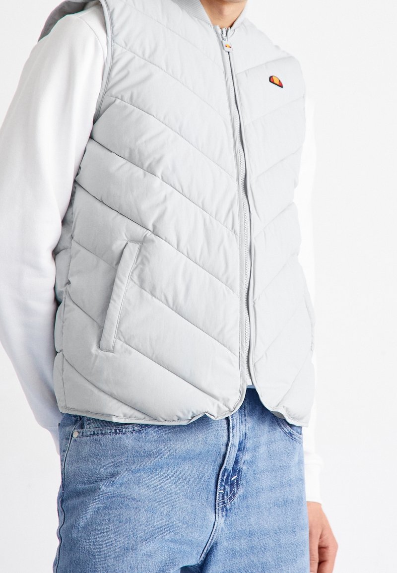 Personne portant un gilet matelassé gris clair avec des coutures diagonales par-dessus une chemise blanche à manches longues et un jean bleu, les mains dans les poches.