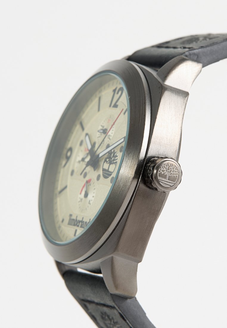 Reloj de acero inoxidable plateado con esfera beige, correa de cuero negro y detalle del logo en la corona. Presenta marcadores de hora en negrita y subesferas.