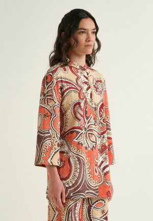 Woman wearing a long-sleeve, buttoned tunic and matching pants with an orange, red, and cream paisley pattern.