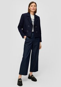 Ciemnoniebieska krótka kurtka z podwójnym zapięciem na guziki, zestawiona z szerokimi spodniami dżinsowymi i czarnymi loafersami z akcentem łańcuszka.