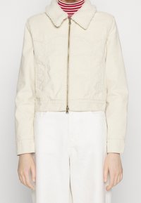 Avskuren, beige jacka med en mjuk, syntetisk pälskrage och dragkedjestängning; har sidofickor och en enfärgad textur. Bärs över en randig topp.