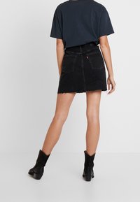 Osoba v černé džínové minisukni s lemem, černých kotníkových botách a tmavém tričku s krátkým rukávem, stojící na pozadí bílé plochy.