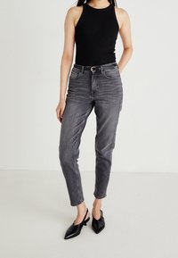 Svart ärmlös bodysuit tillsammans med högmidjade, medelgrå jeans. Jeansen har framfickor och ett klassiskt bälte, avslutat med spetsiga svarta skor.