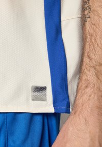 T-shirt de sport blanche avec des accents bleus, arborant une étiquette à l'ourlet. Le t-shirt a une surface texturée et est associé à un short bleu.