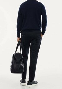 Tmavě modrý svetr, černé kalhoty a černé tenisky zobrazené zezadu, držící koženou cestovní tašku s kontrastním zipem.