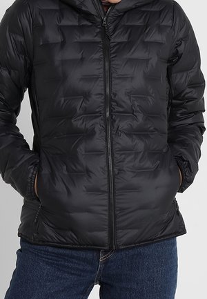 Personne portant une veste matelassée noire zippée, les mains dans les poches latérales et un jean bleu visible de la taille vers le bas.