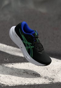 Μαύρο αθλητικό παπούτσι τρεξίματος με πλέγμα στην επάνω πλευρά, που διαθέτει πράσινες και μπλε λεπτομέρειες, μαξιλαρωτή σόλα και κομψό σχεδιασμό. Οι κορδόνες περνούν μέσα από θηλιές.