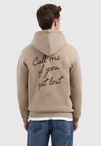 Man in een beige hoodie met de capuchon over zijn hoofd en de tekst "Bel me als je verloren bent" gedrukt op de achterkant, gecombineerd met blauwe spijkerbroek.