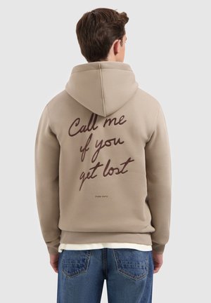 Man in een beige hoodie met de capuchon over zijn hoofd en de tekst "Bel me als je verloren bent" gedrukt op de achterkant, gecombineerd met blauwe spijkerbroek.