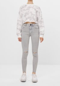 Sudadera cortada tipo tie-dye en gris claro, combinada con jeans ajustados gris claro con roturas en las rodillas, llevados con zapatillas blancas.