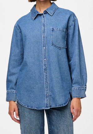 Person iført løs lys blå denim skjorte med knapper og brystlomme samt matchende denim jeans mod hvid baggrund.