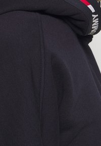 Sudadera con capucha en color azul marino con una textura suave. Presenta una banda de logo en contraste y un bolsillo tipo canguro. El material parece ligero y elástico.