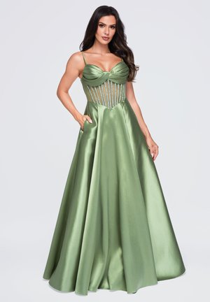 Femme portant une robe longue en satin vert brillant avec fines bretelles et corsage rayé en mesh, debout avec une main dans la poche.