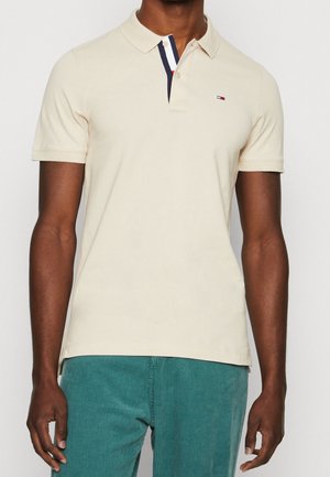 Mand iført beige kortærmet polo med lille logo på venstre bryst og blågrøn fløjlsbukser, stående mod en ensfarvet baggrund.