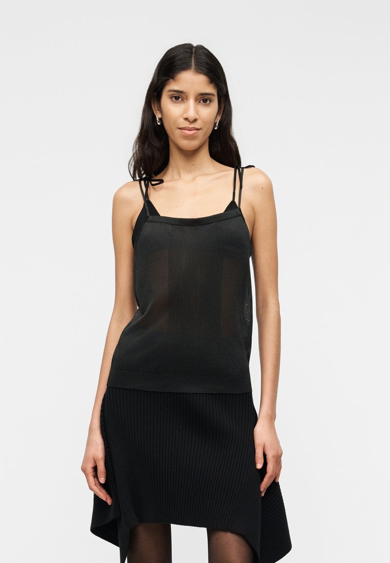 Νεαρή γυναίκα με μακριά σκουρόχρωμα μαλλιά φοράει μαύρο αμάνικο τοπ με λεπτές τιράντες και ασύμμετρη μαύρη φούστα, στέκεται μπροστά σε απλό φόντο.