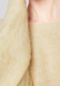 Beigefarbener, flauschiger Strickstoff mit einer weichen Textur. Nahaufnahme eines Kleidungsstücks mit lockerem Gewebe und leichtem Glanz, das um den Hals gewickelt ist.